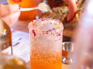 A person garnishing a glass of an orange cocktail drink with ice and a sugar rim, surrounded by bottles, a small jigger, and fresh herbs.